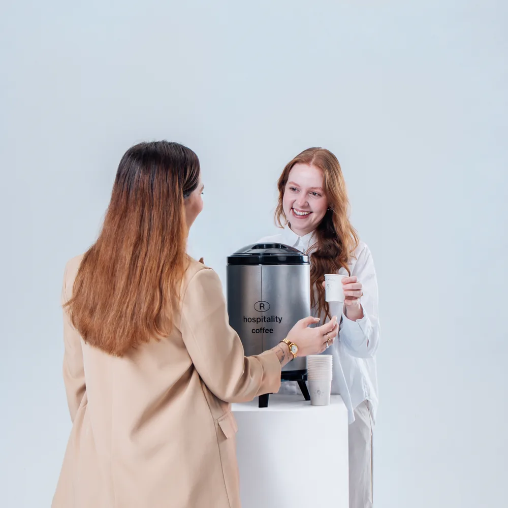 Duas mulheres servem café na térmica, de costas, no estúdio