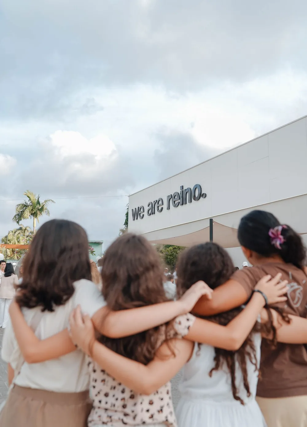 Quatro meninas abraçadas, de costas, olhando o letreiro we are reino