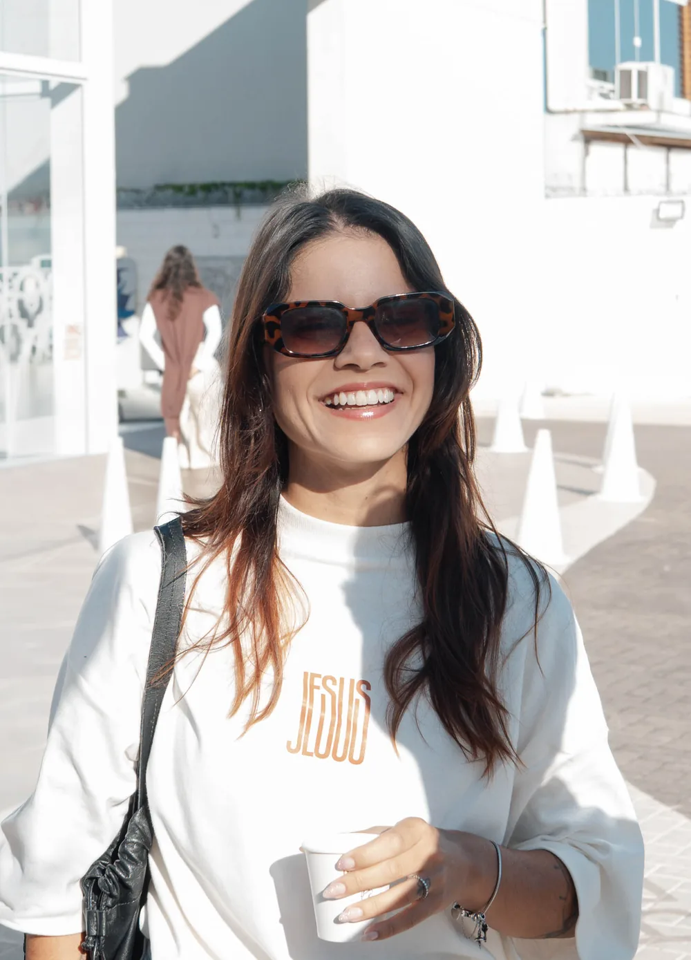 Mulher de óculos escuros e camiseta Jesus sorri com o copo de café